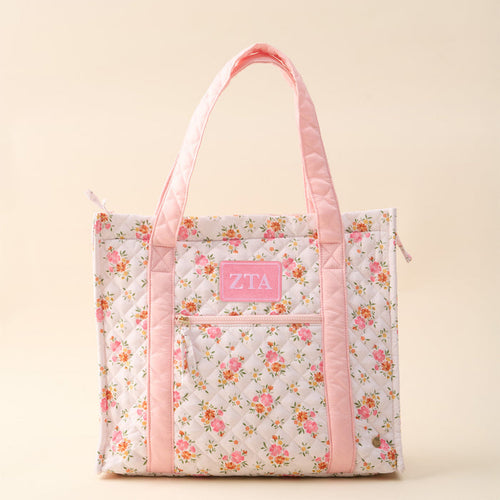 The Darling Effect Zeta Tau Alpha The Everyday Quilted Tote Bag features a soft beige quilted design with pink handles, floral trim, a front pocket, and a pink ZTA patch—perfect for sorority members’ everyday use.